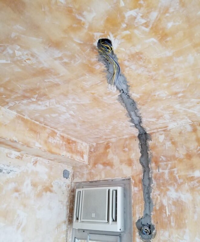 室內天花及牆角剷底後實況,天花及牆面呈橙黃色舊防潮底油底層,可見牆角天花有電線穿出及水泥填縫修補,舊窗口式冷氣機安裝於右下方牆身