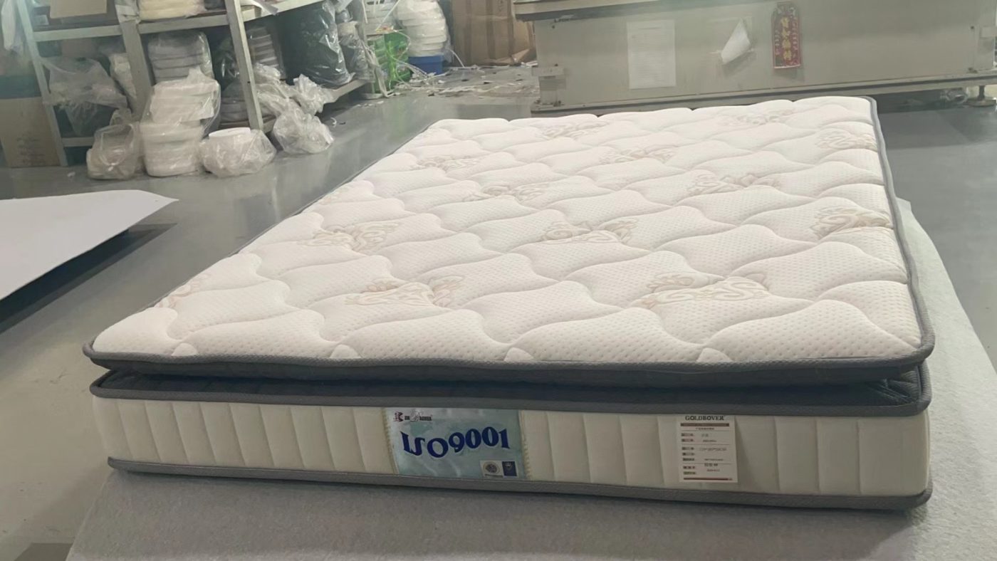工場內一張床褥平放喺工作枱上，白色絎縫布面帶淺色花紋及波浪形起伏，四周配深灰色包邊；米白色側圍有直向縫線，前方附有標籤，後方可見貨架同機器。