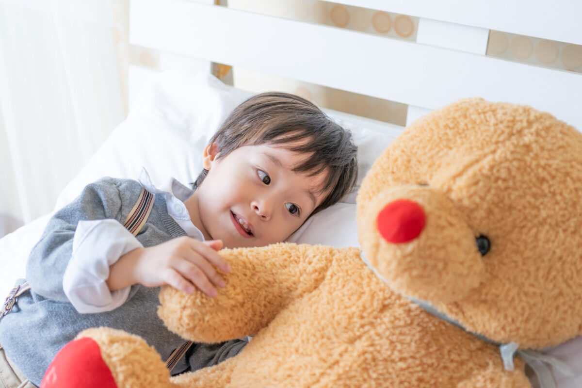 亞洲小男孩側躺於白色床褥上，懷抱米黃色大型泰迪熊，白色床頭板背景，窗簾透進自然光，床鋪整潔乾淨