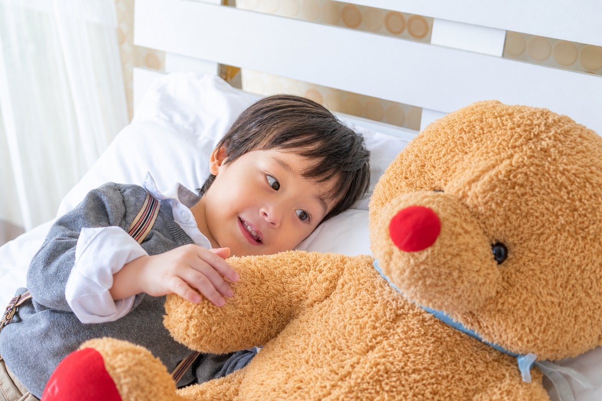 亞洲小男孩側躺於白色床褥上，懷抱米黃色大型泰迪熊，白色床頭板背景，窗簾透進自然光，床鋪整潔乾淨
