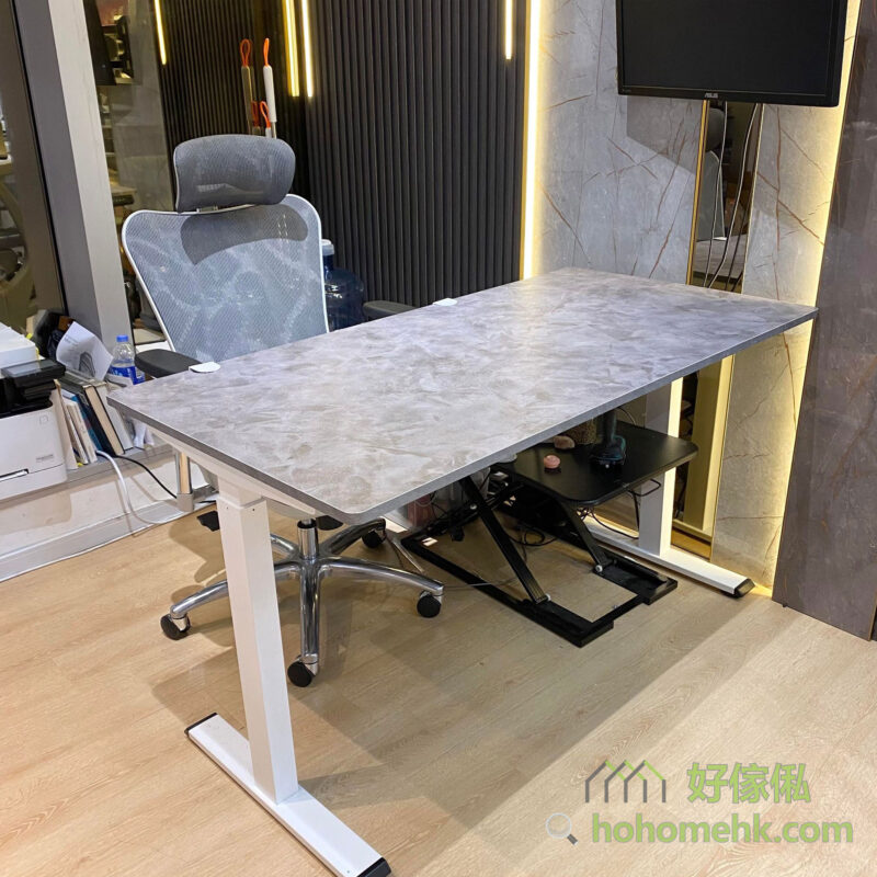 Morton電動升降枱展示間側面實物照片，灰色水泥石紋矩形枱面配白色T字形升降鋼腳，旁邊放有灰色網背人體工學辦公椅，枱下可見黑色腳踏架，背景為深色吸音板牆身及金色裝飾邊框，右側有電視螢幕