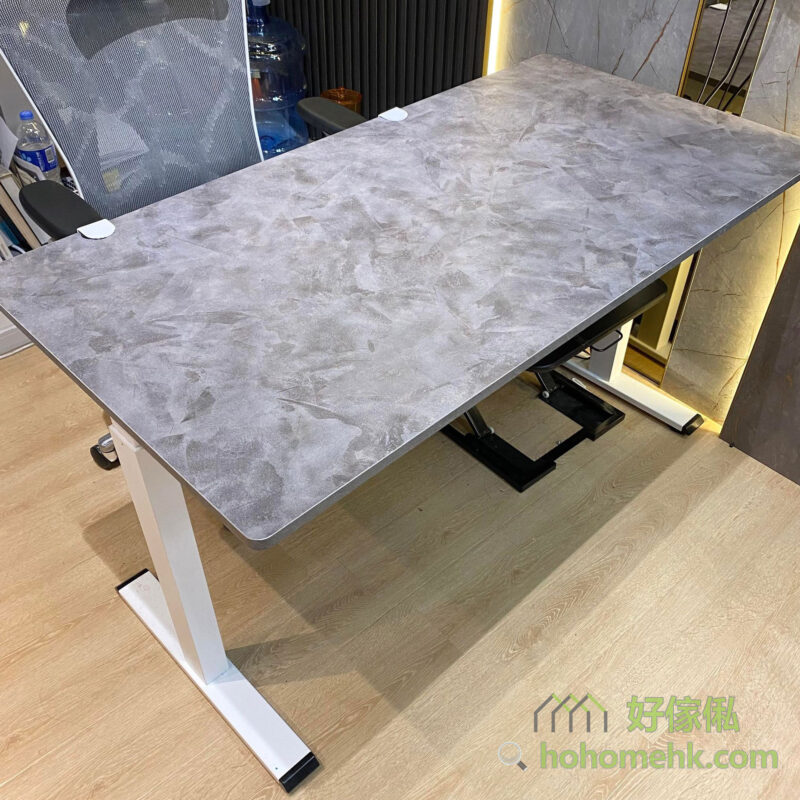 Morton電動升降枱展示間低角度實物照片，灰色水泥石紋矩形大枱面配白色T字形鋼腳，枱面四角有白色保護角墊，枱面紋理清晰可見石紋肌理，背景為展示間空間，右側見網背辦公椅