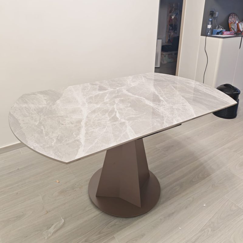 室內木紋地板上擺放伸展後的岩板餐枱，灰色雲石紋亮面枱面形成長形圓角輪廓，中央配啡灰色幾何座地枱腳，旁邊是白牆及儲物櫃。