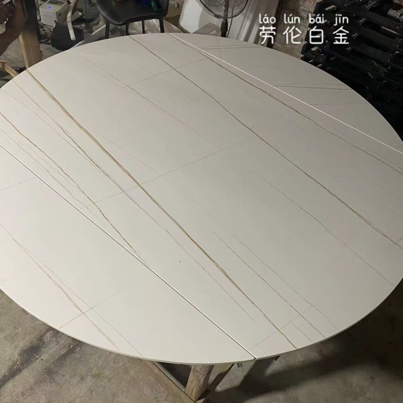 工場內近距離拍攝圓形岩板餐枱，啞光暖白色枱面配幼細金啡色斜紋，多組拼接線橫跨枱面，餐枱放於木架上，後方可見座椅及風扇。