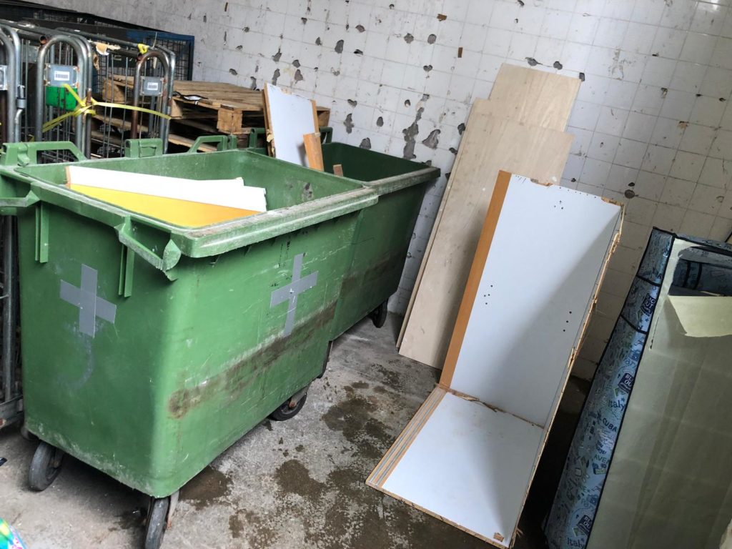室內廢物存放區,綠色大型滾輪環保垃圾箱旁靠著多塊白色夾板及木色板材,牆壁磁磚剝落,傢俬廢料整齊靠牆放置