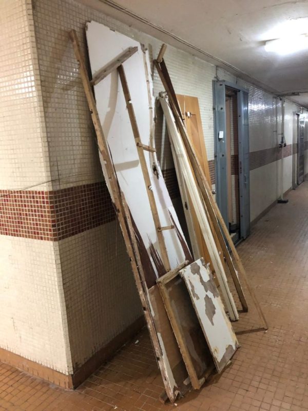 住宅大廈走廊磁磚牆邊,多塊拆除後的白色夾板及木框架靠牆豎放,走廊盡頭可見大廈門口及電燈,等待運載的清拆廢料