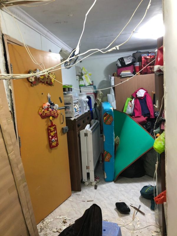舊式住宅房間清拆中,牆壁局部拆除,天花橫跨白色電線,房內堆滿衣物袋、電暖爐、行李等舊物,地上散落碎料