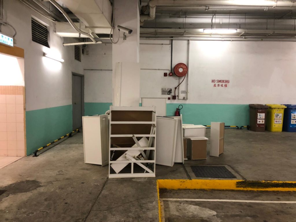停車場內白色牆身前,多件白色系傢俬組件拆散後堆放地上,包括書架架身、抽屜側板及白色板材,場地有紅色消防裝置及英文禁煙告示