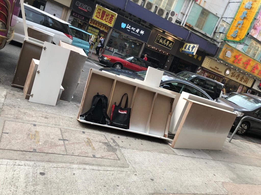 香港旺區街道行人路上,多件木色夾板傢俬組件橫放地上,背景為香港舊式商鋪招牌及街道車輛,師傅工具袋放於組件旁