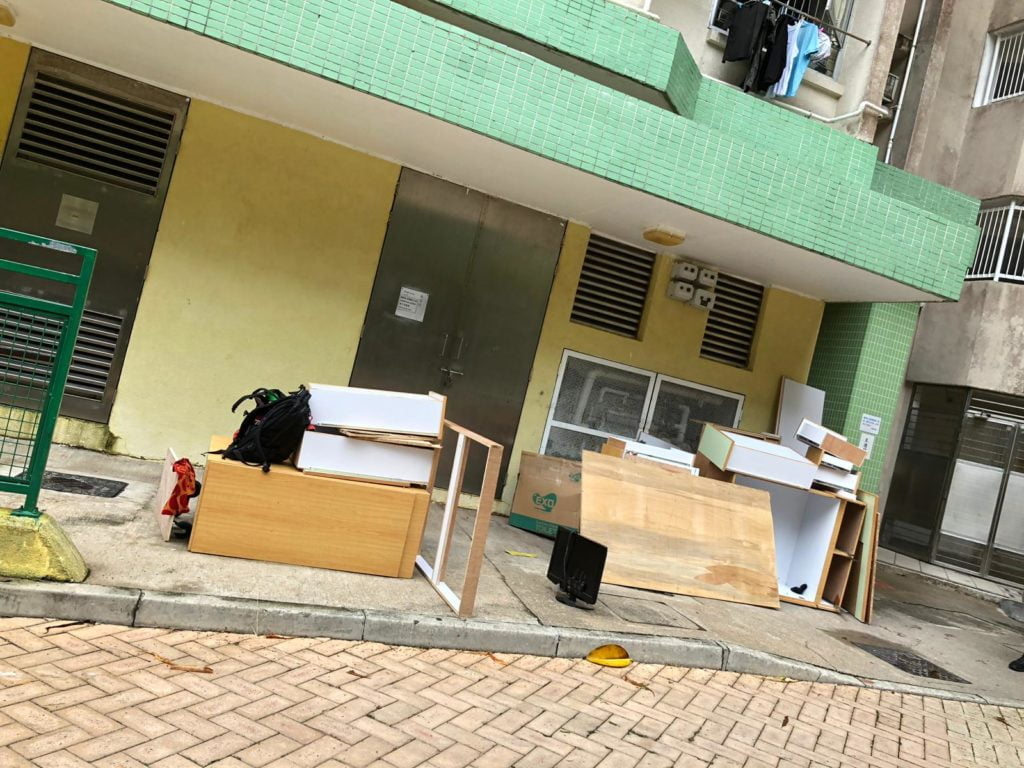 香港公共屋邨地下外牆旁,多件白色及木色夾板傢俬板材整齊疊放於行人路上,背景為黃綠色磁磚外牆及大廈出入口