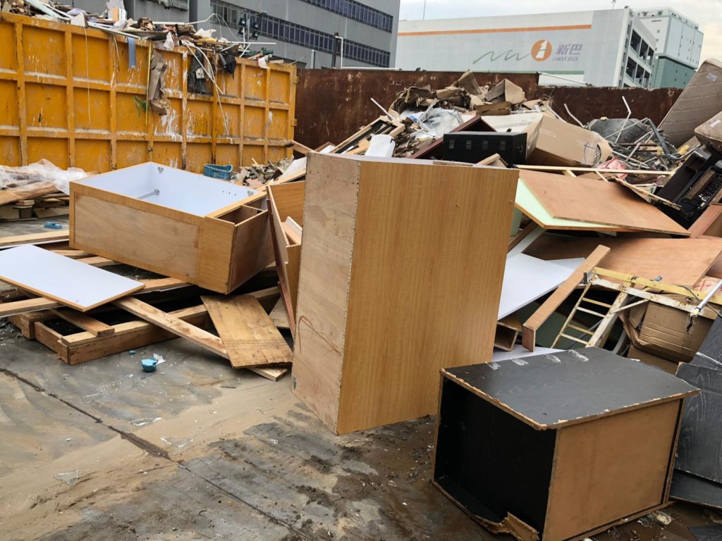 廢物收集站戶外場地,大量拆解後的傢俬板材、衣櫃組件及木材廢料堆放於大型黃色鐵箱旁,遠處可見商業大廈,天色陰沉