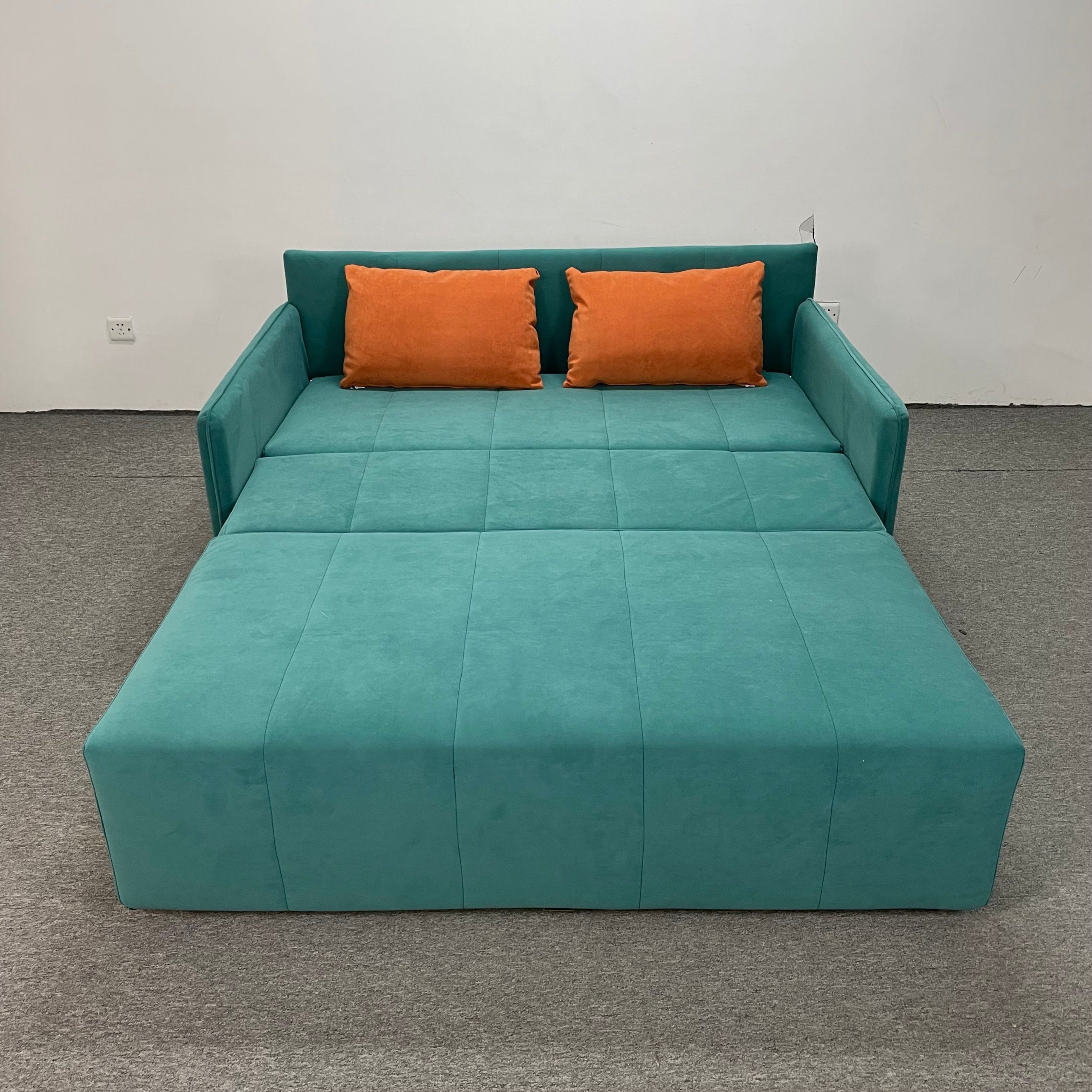 孔雀綠絨布Takumi梳化床展開成床狀態正面圖，背靠板立起兩側作床頭，床面平整寬闊分三格，配兩個橙色抱枕橫放於背靠前，白牆背景，灰色地毯