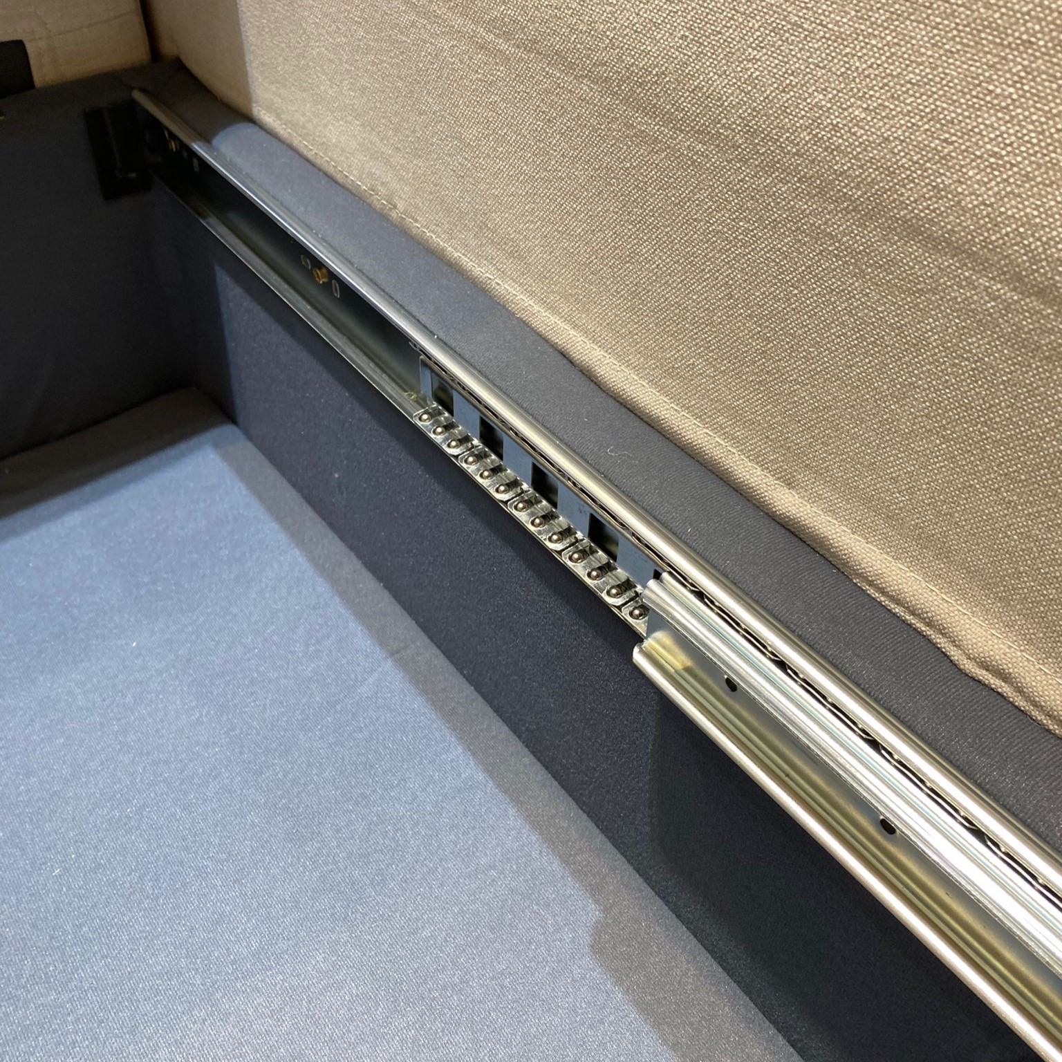 梳化床底部走珠不銹鋼軌道近距離特寫，銀色雙軌道清晰可見滾珠結構，旁邊為深灰色床箱側板，底部露出藍色床褥面，由側面往下拍攝