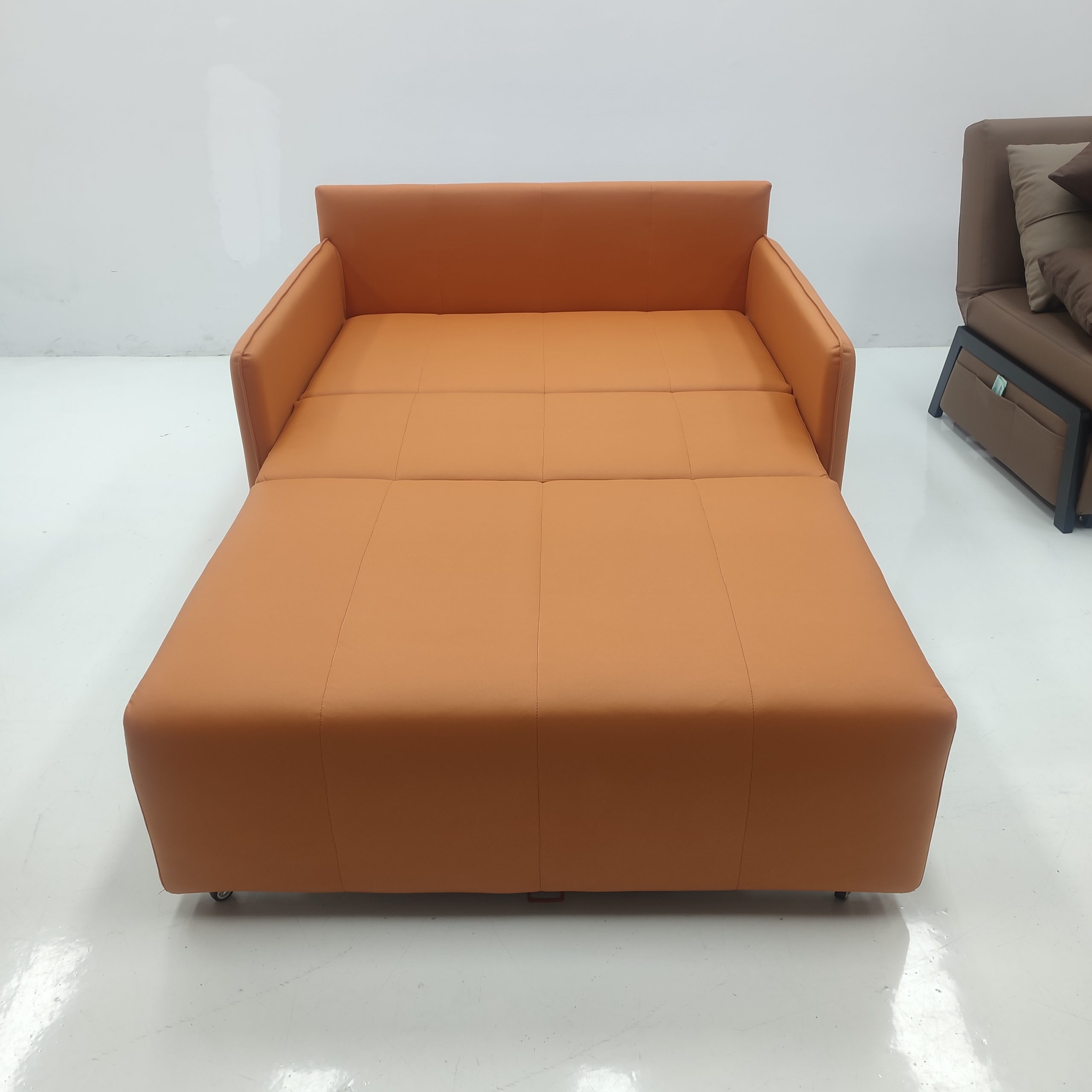 Takumi 6079款梳化床以橙色科技布包覆，圖為展開成雙人床鋪狀態，正面角度可見平整的分格縫線床面及高扶手靠背，底部裝有小滑輪方便移動，旁邊有另一張啡色梳化床作參考
