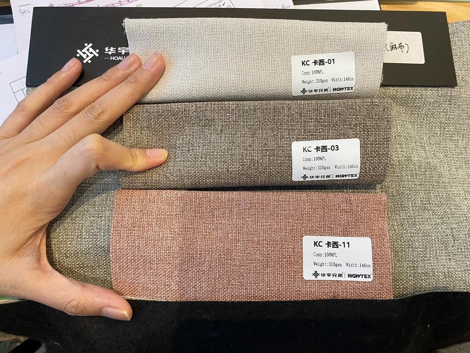 門市KC卡西系列布料色板三塊並排，由上至下淺灰色KC-01、中灰色KC-03及玫瑰粉KC-11，布面呈細紋麻料質感，標籤標示100%NPL成分，旁有人手作比例參考