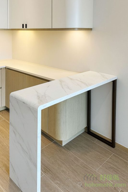 在開放式廚房與客廳之間，安裝一張訂造的高身吧枱，實現了空間分隔的功能，還提供了額外的備餐枱面。吧枱表面以白雲石紋飾面設計，枱邊做了圓邊設計，既耐用又易於清潔。此外，吧枱還能兼作餐枱，讓小空間發揮多功能用途。